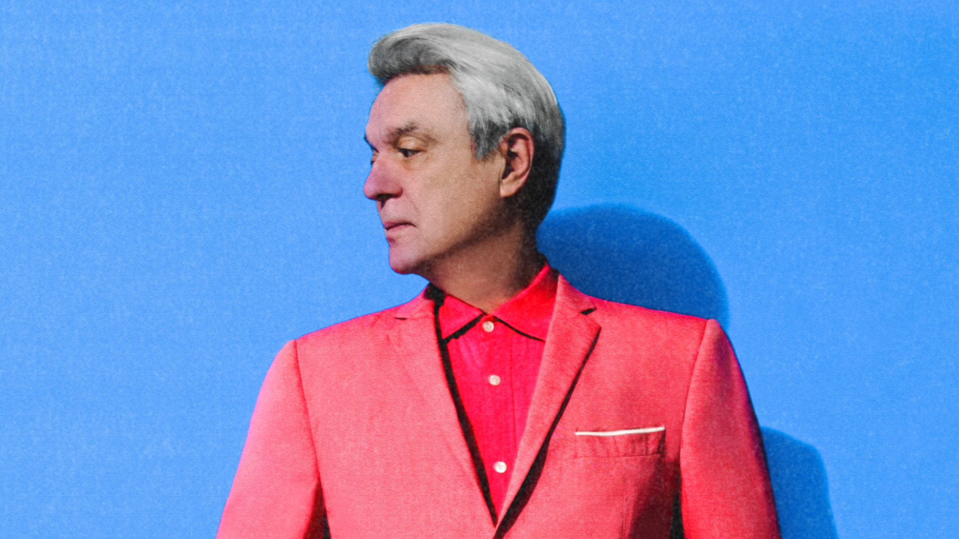 David Byrne in a red suit and shirt on a blue background