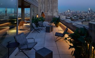 View of a rooftop terrace at The Robey, Chicago, US featuring tiled flooring, grey chairs, grey block tables and green plants in pots. There are also views of surrounding buildings in the evening