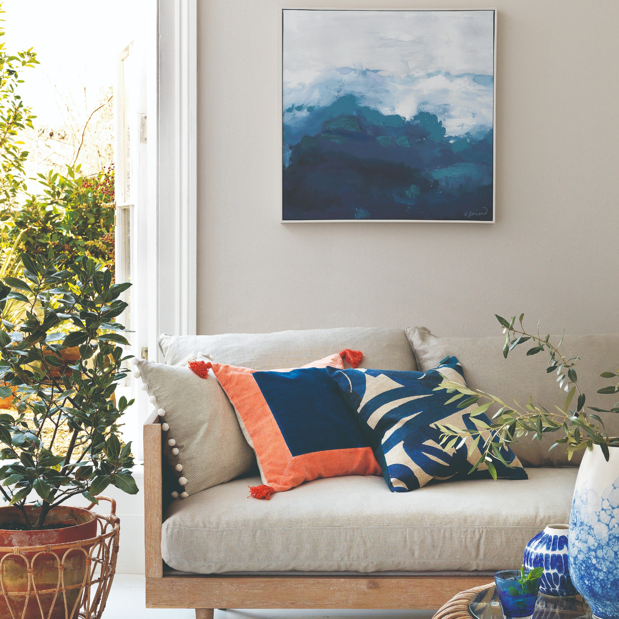 A bright living room with a beige wooden-frame sofa and an abstract painting hung on the wall behind