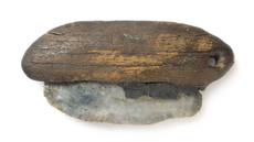A whitish stone tool is stuck into a piece of brown wood with greyish tar. There is a hole drilled into the wood.