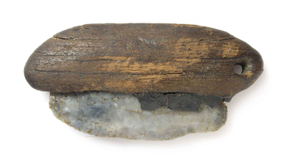 A whitish stone tool is stuck into a piece of brown wood with greyish tar. There is a hole drilled into the wood.