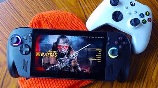 ROG Xbox Ally X sitting on orange beanie next to white Xbox wireless controller with Fallout New Vegas main menu on screen.