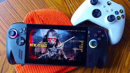 ROG Xbox Ally X sitting on orange beanie next to white Xbox wireless controller with Fallout New Vegas main menu on screen.
