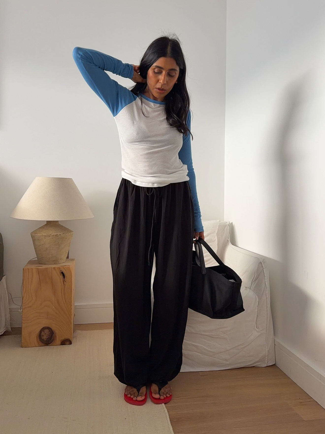 British style influencer Monikh Dale poses in her London bedroom wearing a contrast raglan tee with blue sleeve, black balloon pants, a black tote bag, and red The Row flip-flop sandals.