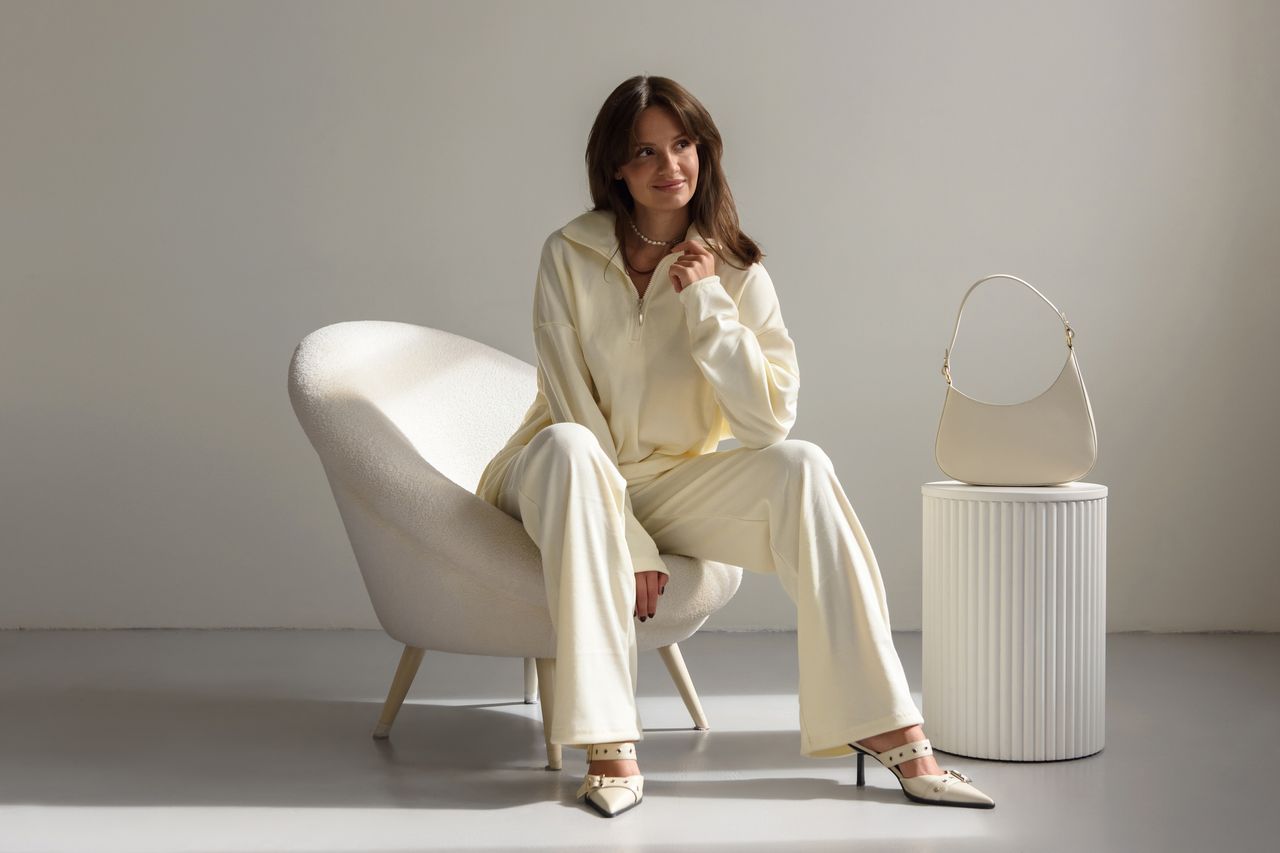 A female model wearing a white long-sleeve trouser set while sitting on a white chair, with a white bag next to her. 