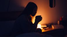 Woman holding cup of tea in the middle of the night, lit by side table lamp