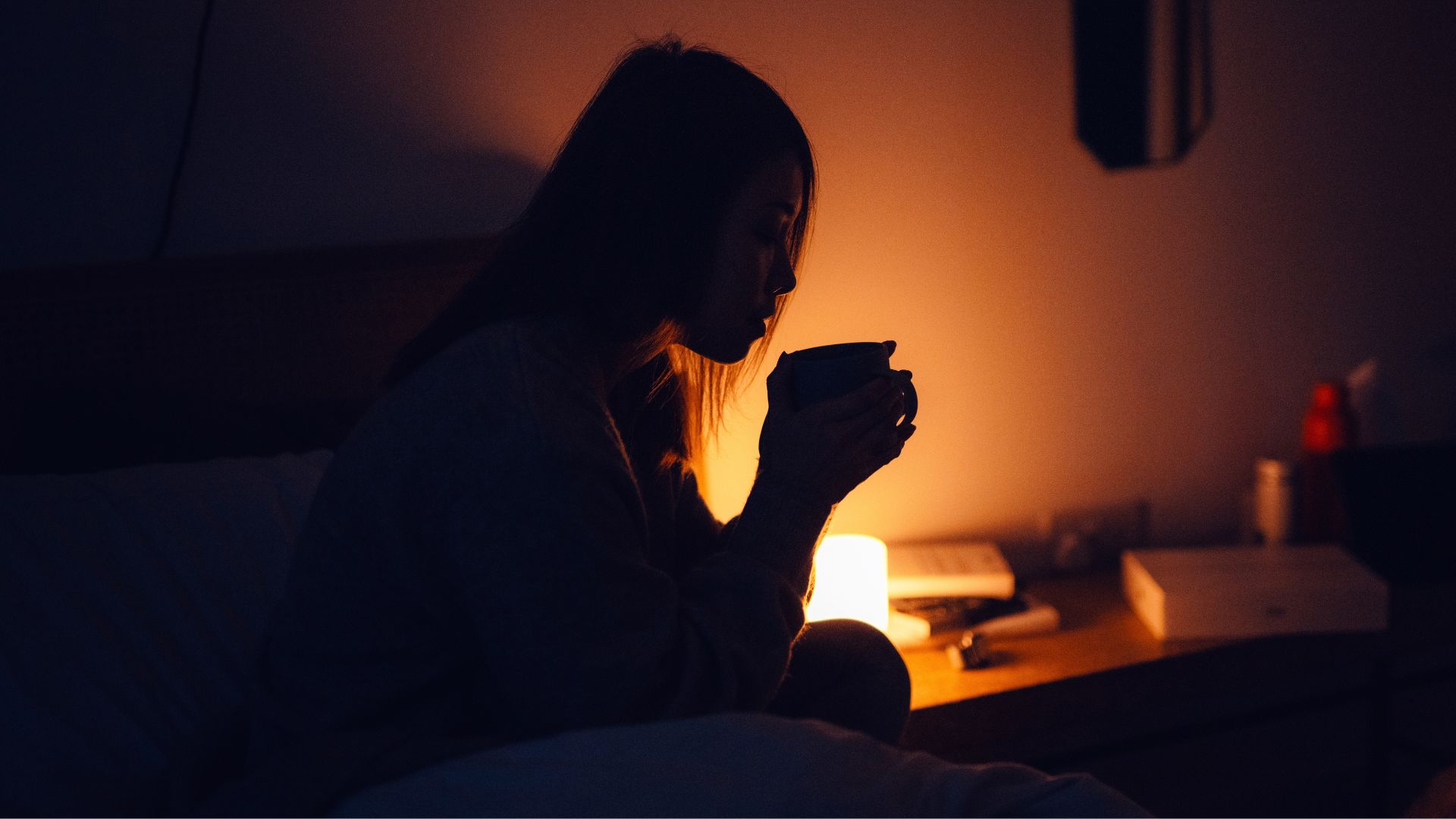 Woman holding cup of tea in the middle of the night, lit by side table lamp