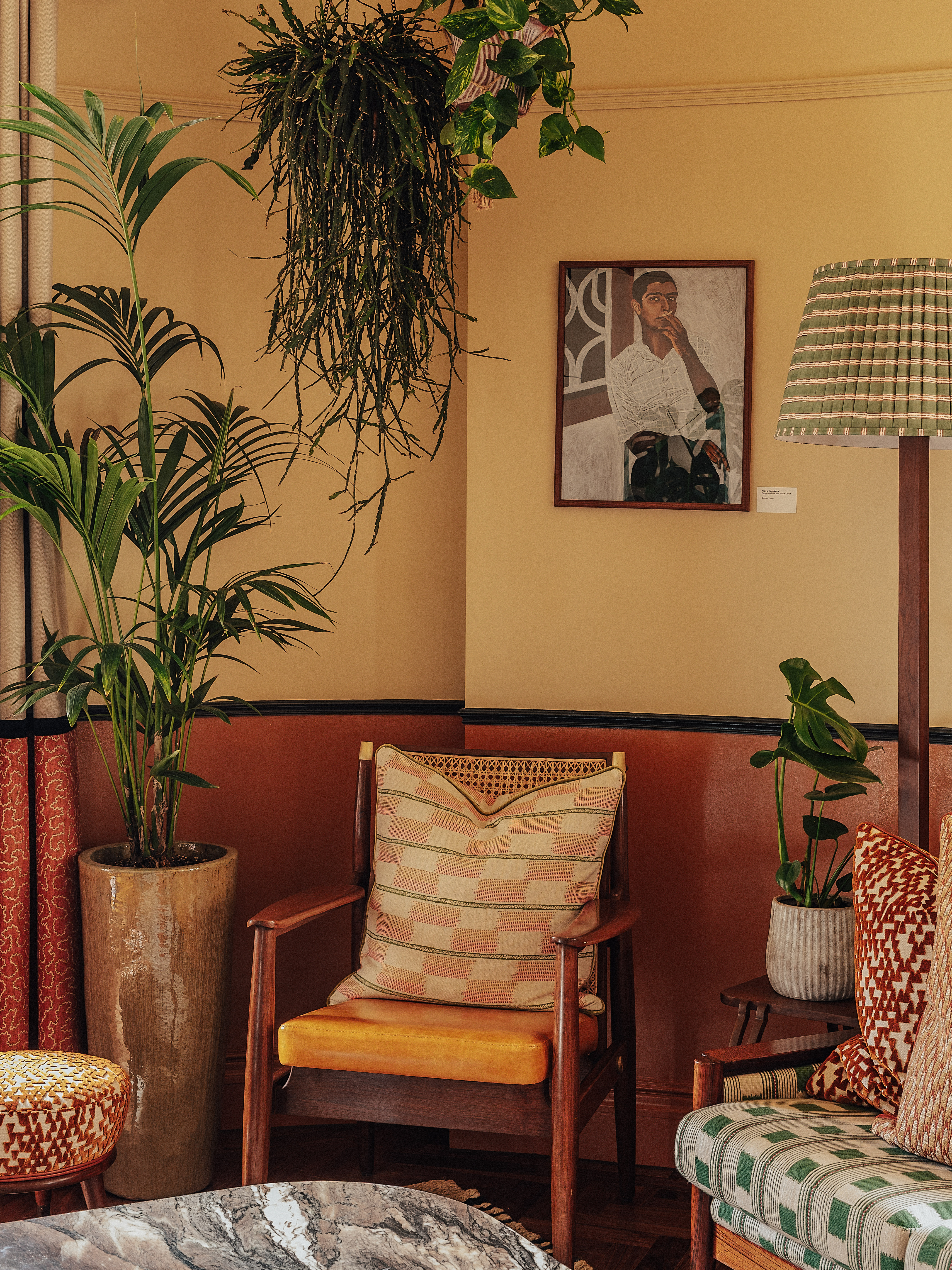 A vibrantly decorated hotel suite inspired by Bombay residences features buttery or tile blue walls adorned with original portraiture artworks, bespoke retro-fueled furnishings in wood, leather, and patterned fabric, and thriving plants.