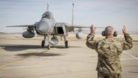 A large gray plane rolls on a runway with a man wearing army fatigues waving it forward