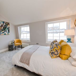 Stylish bedroom with white linen and mustard pillows with a mustard chair in the corner on a luxurious grey carpet