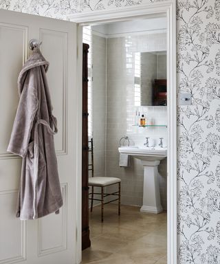 A gray bathrobe hanging on the back of an open white bathroom door. Gray flower patterned wallpaper. A white tiled bathroom viewed through the doorway with a pedestal sink and a wooden chair