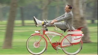 Pee-Wee Herman riding his bike in a park with his feet on the handlebars.