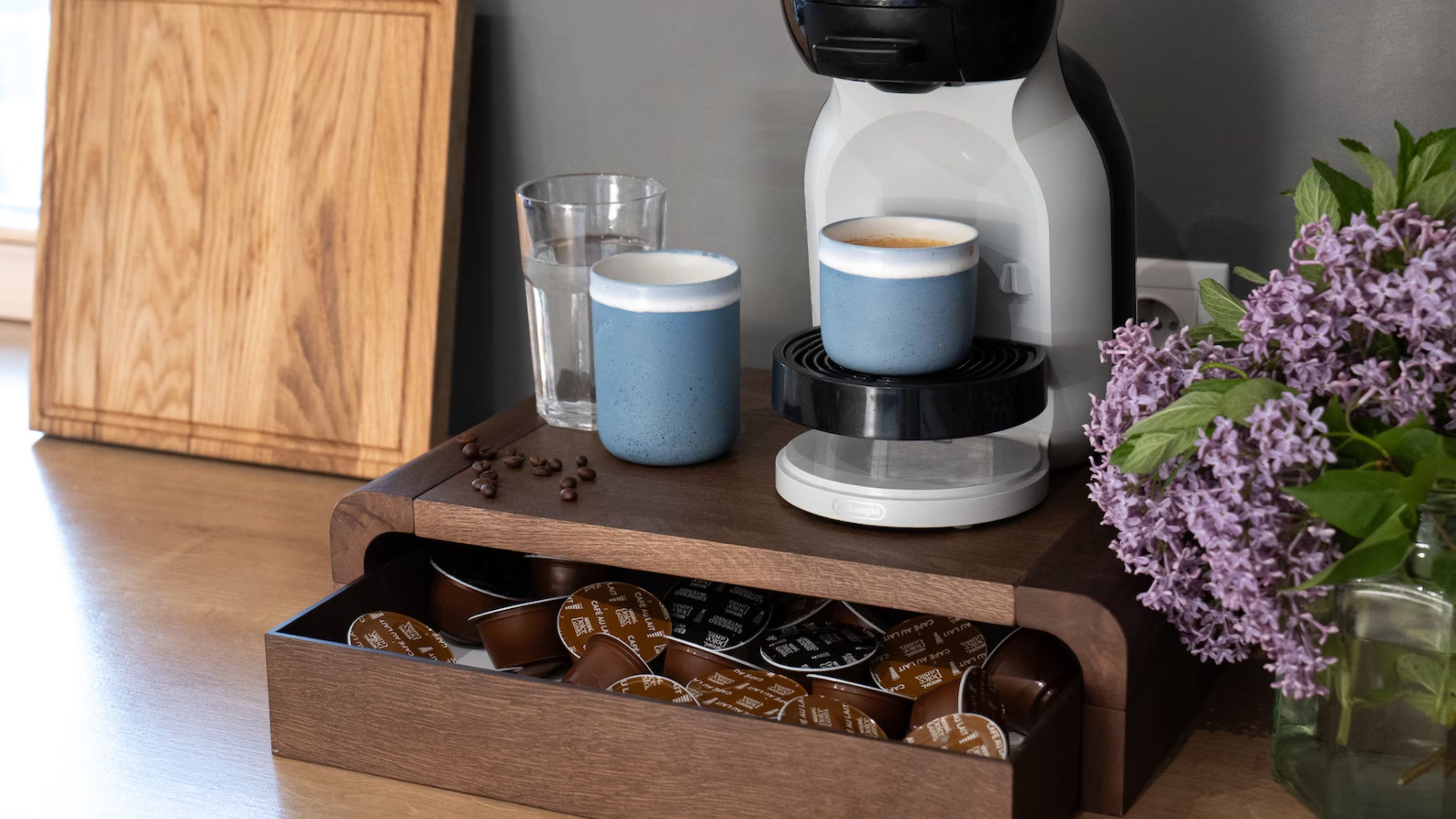 Image of a dark wooden coffee machine stand with a drawer filled with coffee pods. There is a coffee pod machine on the stand and a couple of mugs of coffee.