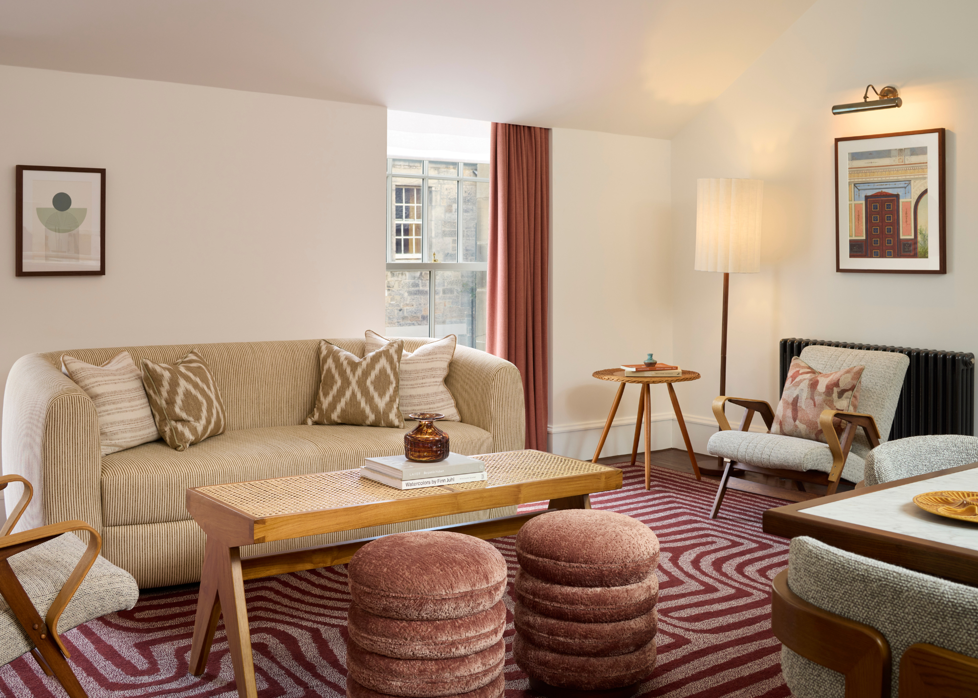 A bright living room with stacked dusty rose pouffes, a wood coffee table and side table, a paper floor lamp, hanging paintings with display lights, a burgundy and pink swirl area rug, and gray armchairs by a stripey couch