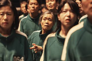 An elderly woman (Kang Ae-shim, center) stands among a crowd of people, all in matching green tracksuits, in 'Squid Game' season 2.