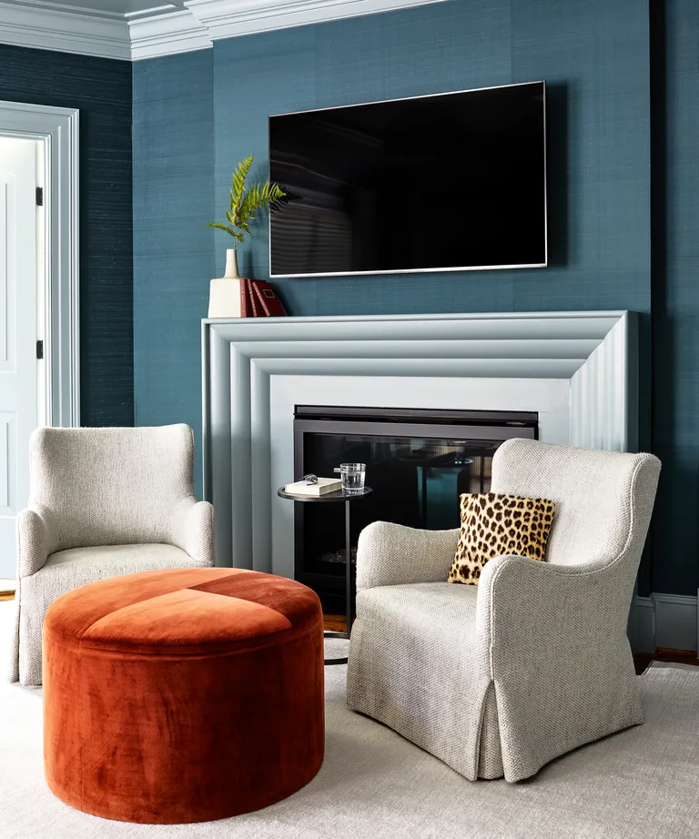 A bedroom TV idea with wall-mounted television on blue wallpaper, over a white fireplace with white armchairs and orange ottoman