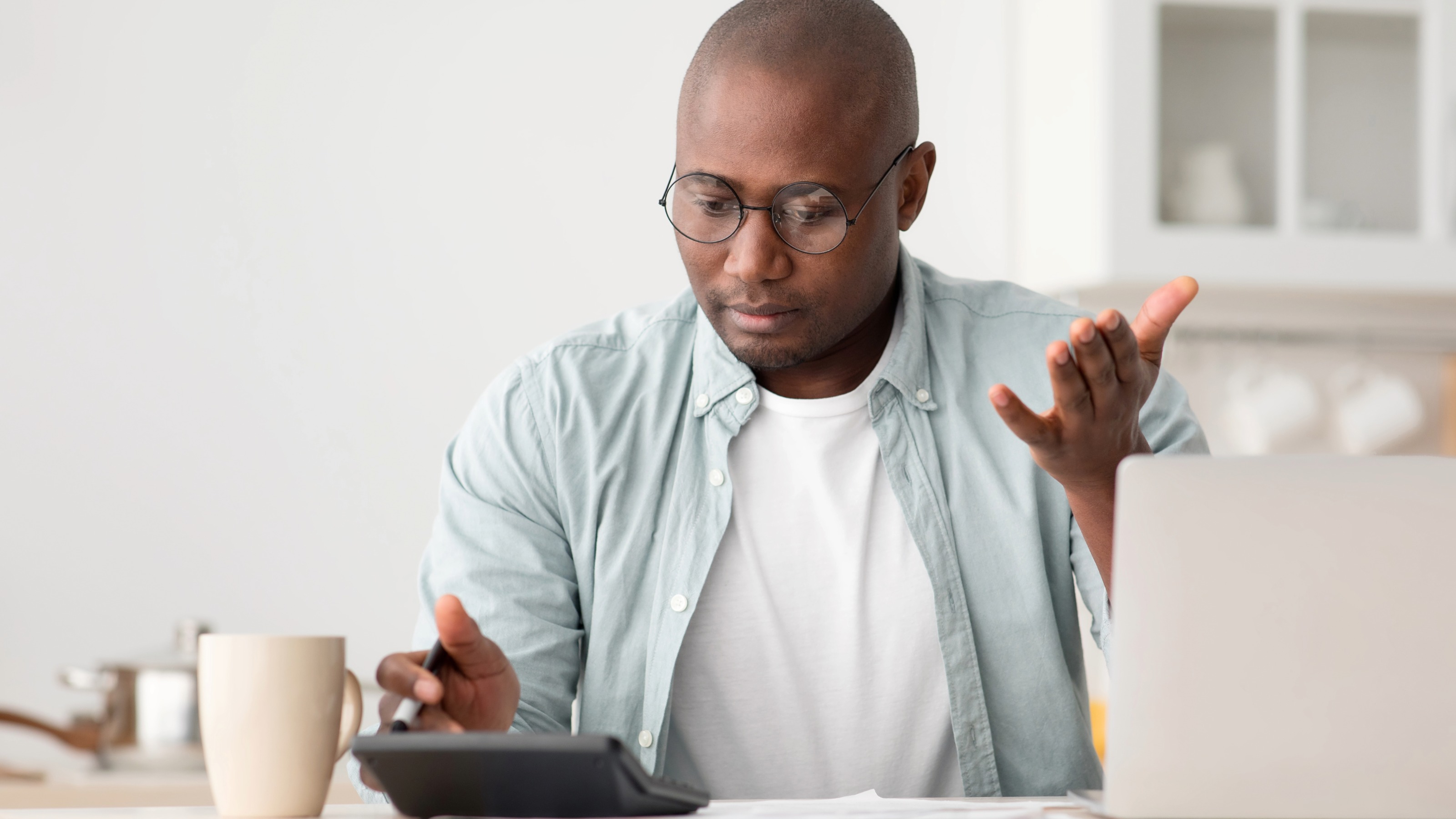Shocked man using calculator and laptop, sitting in the kitchen at home
