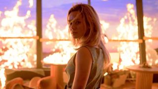 A woman looks behind her with her hair in her face while a room burns behind her