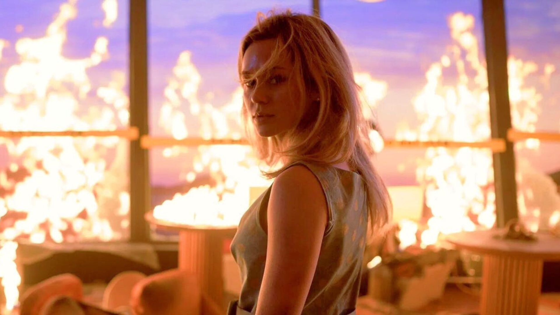 A woman looks behind her with her hair in her face while a room burns behind her