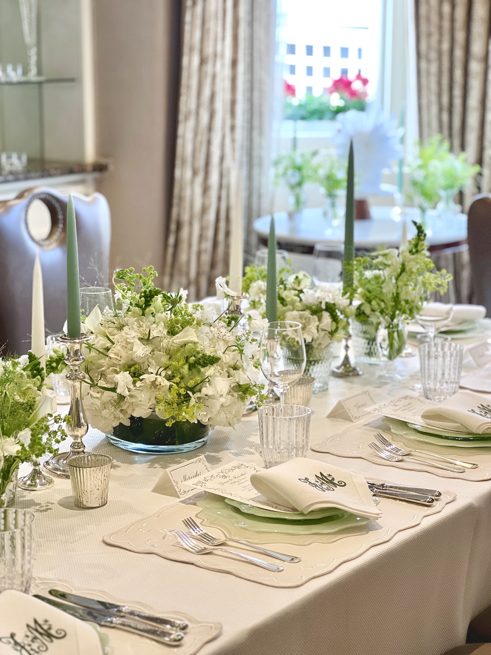 A sophisticated dining table dressed with a cream tablecloth and place settings, with light/lime green plates and embroidered napkins on top. There are bouquet centre pieces, in between silver candlesticks with green and white candles to match the theme.