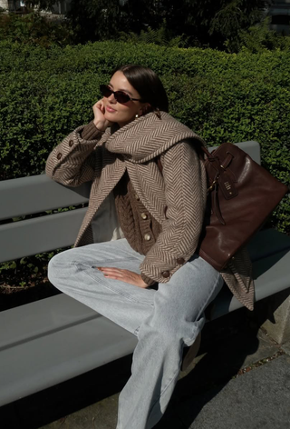 what to wear in 30-degree temperatures shown in a photo of a woman sitting on a bench wearing a herringbone print scarf coat over a chunky brown knit cardigan with light wash jeans, brown ankle boots, and a large miu miu handbag