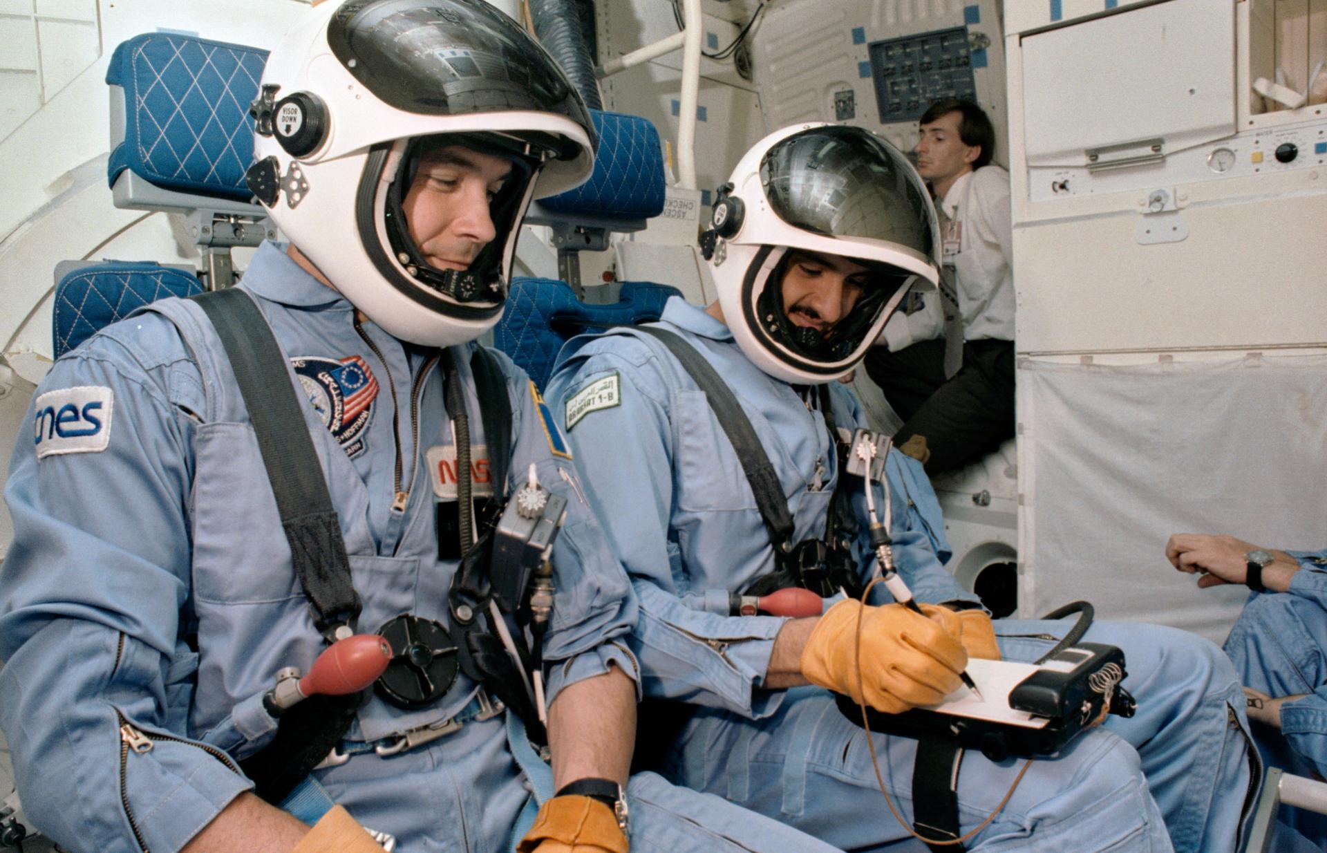 Two astronauts in flight suits and helmets fasten their seat belts on a space shuttle trainer.The one on the right writes on the notepad