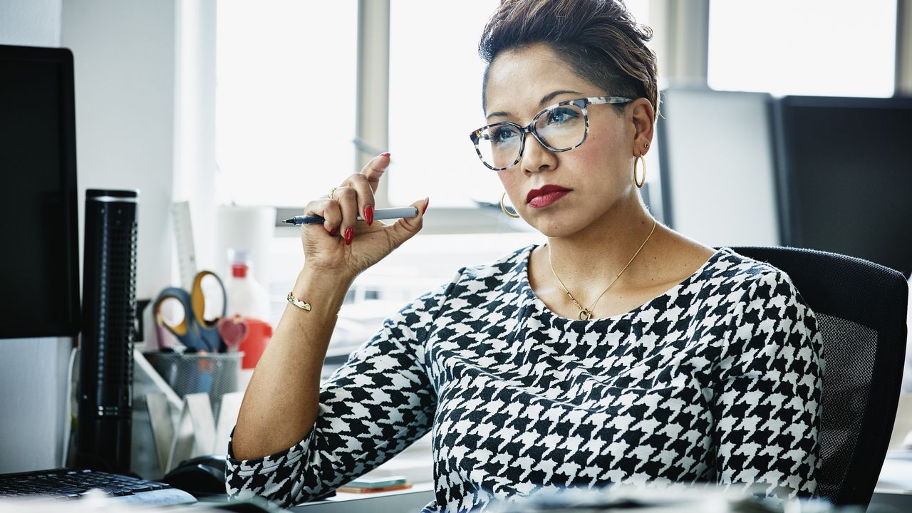 A businesswoman with a serious look on her face sits at her desk in an office.