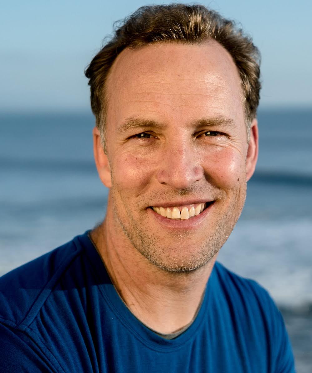 Head and shoulders of man smiling at camera. He wears a blue T-shirt and the sea is in the background