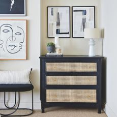 Black chest of drawers with rattan inserts against a white painted wall with artwork on the walls around it