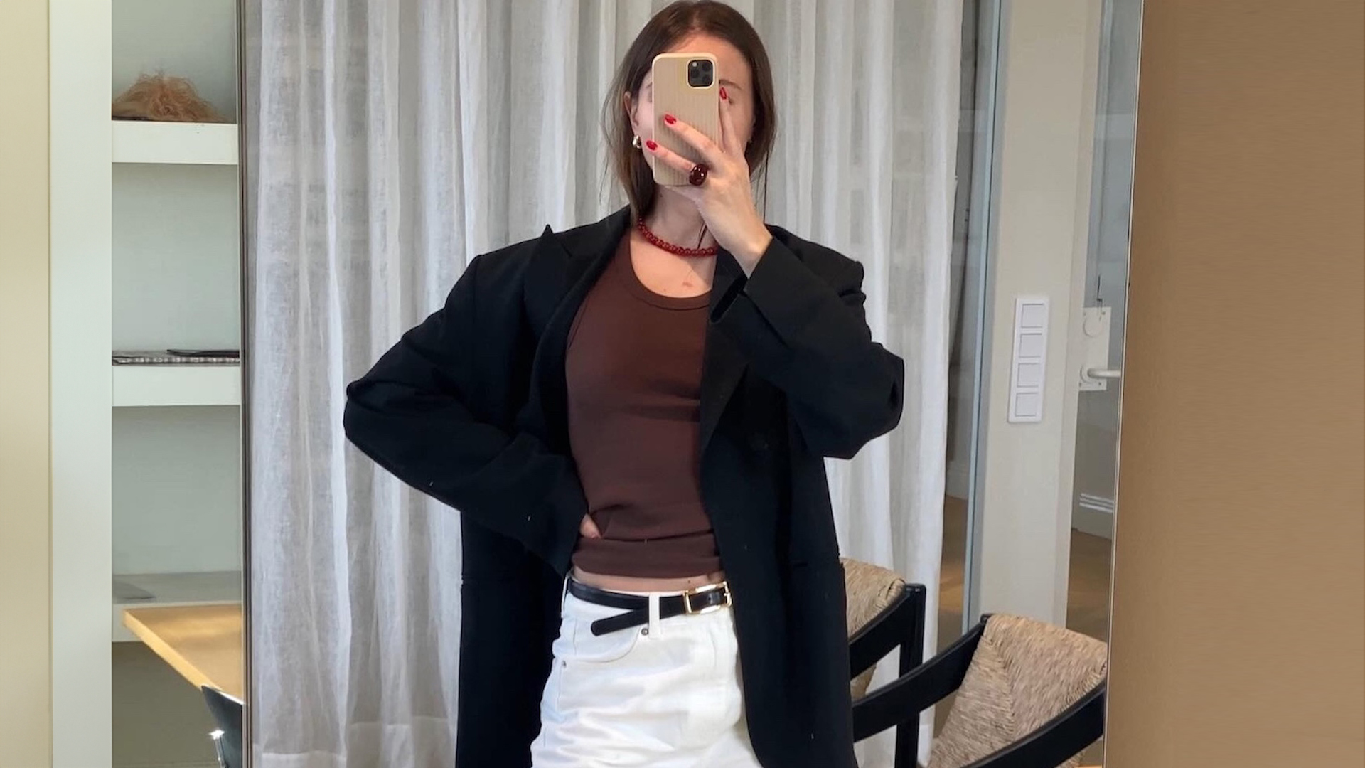 Swedish fashion pr and influencer Johanna Lager poses for a mirror selfie wearing a red beaded necklace, brown T-shirt, oversize black blazer, thin black belt, and white jeans