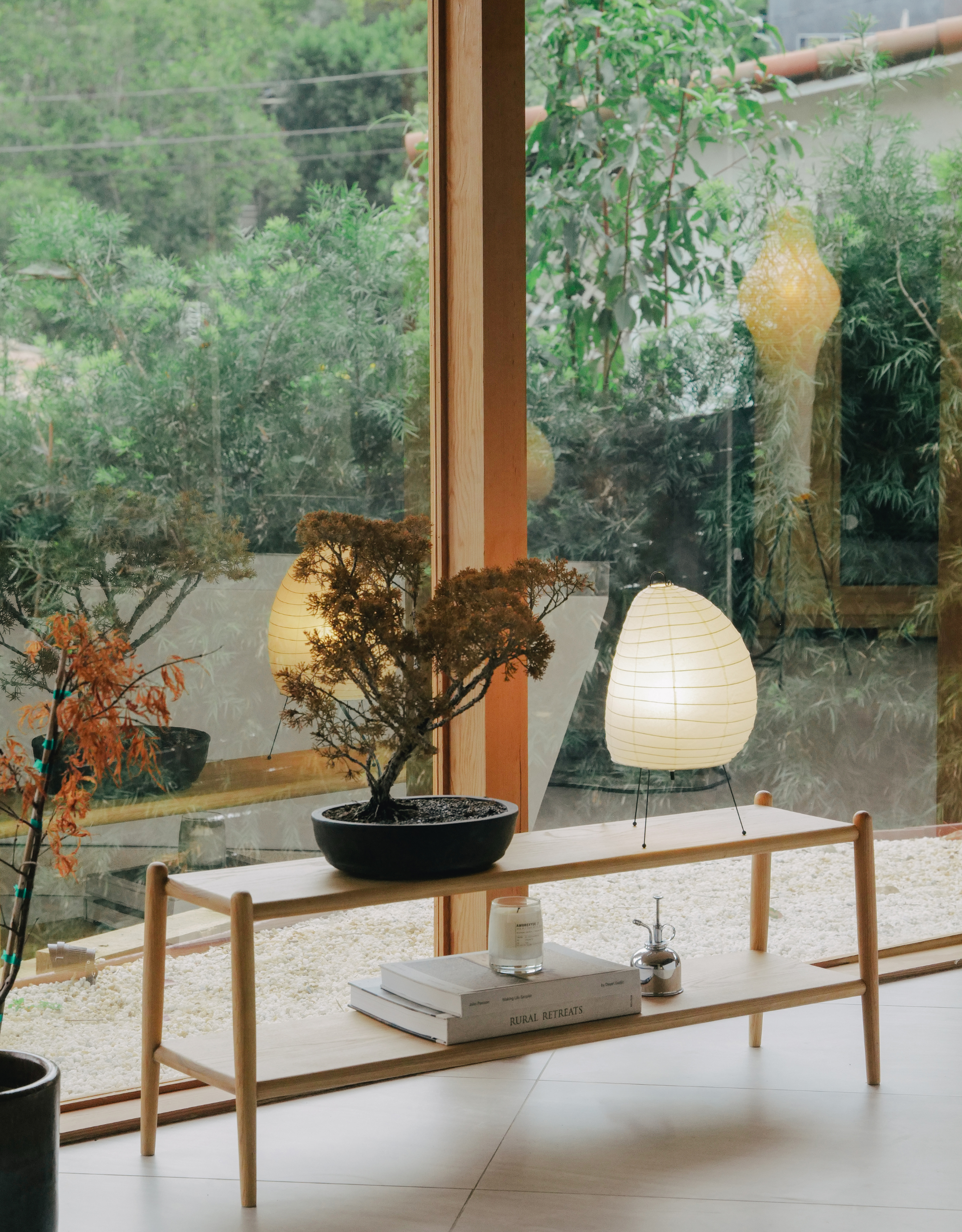 A living room beside a garden with a wood stool, a potted tree, a rice paper table lamp, and a candle