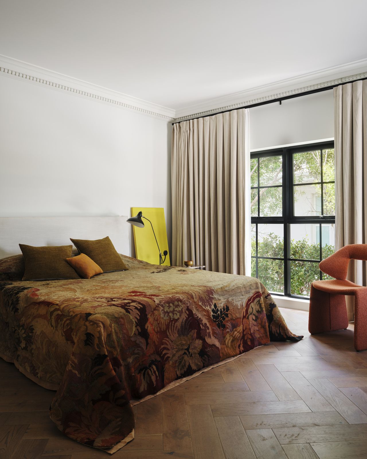 modern bedroom with timber parquetry floors, an orange armchair, windows with beige curtains, a yellow artwork on a bedside table, and a bed with a tapestry bedcover