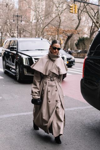 A woman at New York Fashion Week wearing a cape trench coat and black sunglasses walking in the street