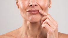 a woman's lower face pictured close up, she is touching her lip and wearing pearl earrings