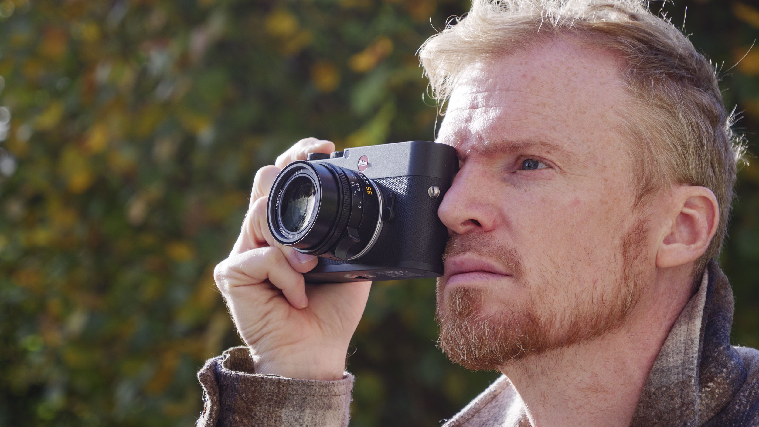 Man holding the Leica M EV1 camera's viewfinder up to their eye, autumn leaf backdrop