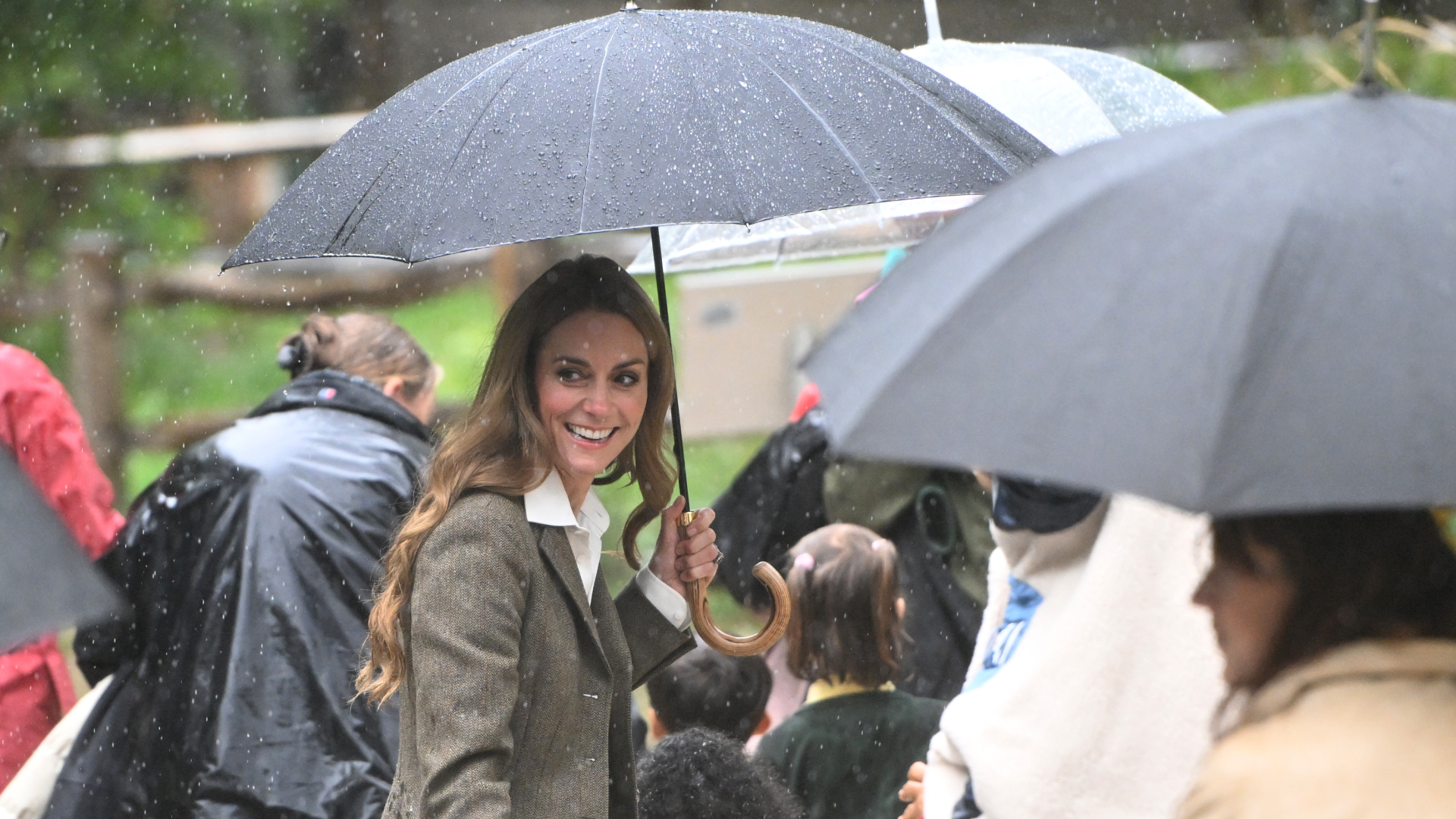 Kate Middleton holding a black umbrella in the rain surrounded by people