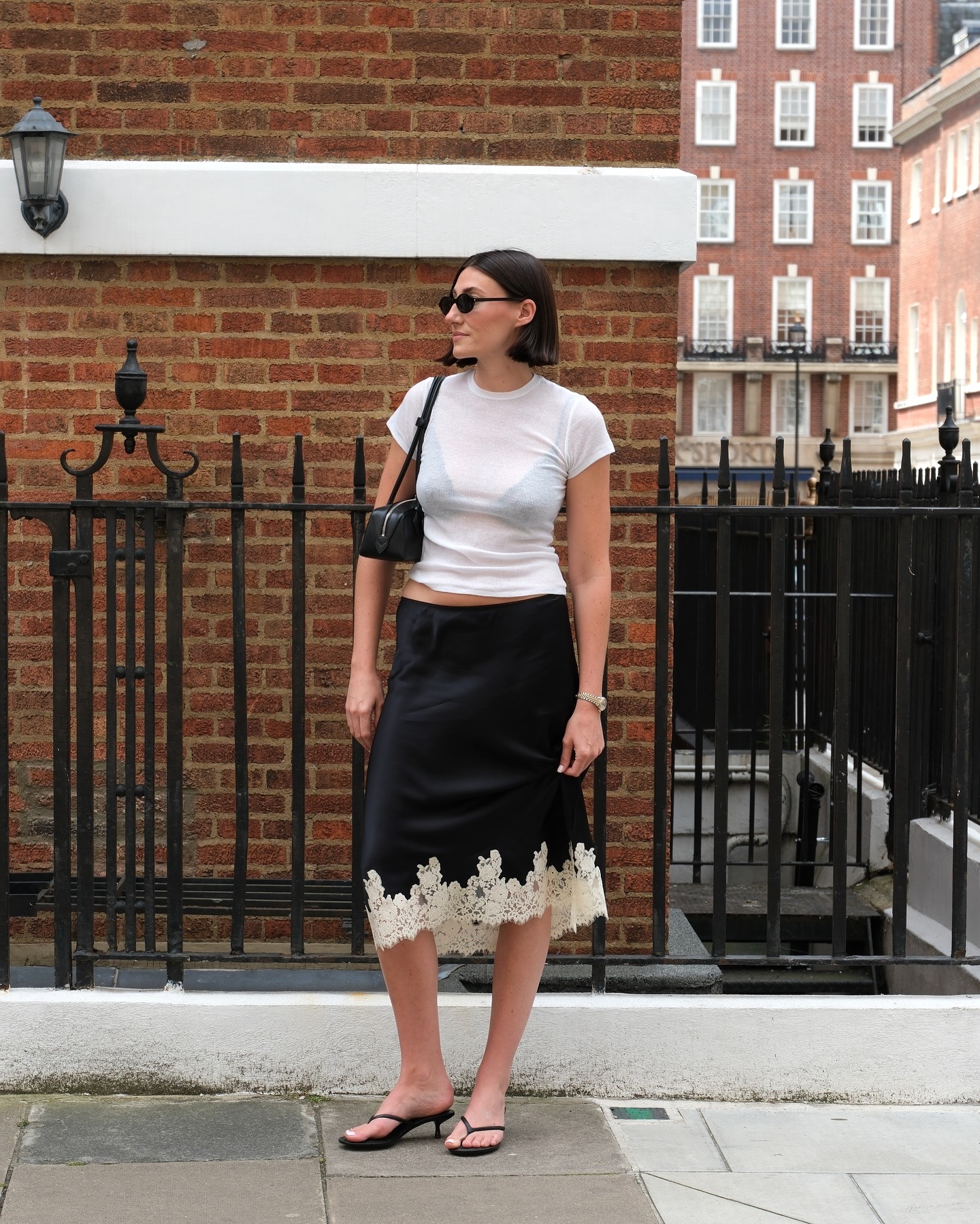 British influencer Sarah Louise Blythe poses on a London sidewalk wearing black oval sunglasses, a sheer white T-shirt, black East-West shoulder bag, a black slip skirt with a white contrast lace hem, and black flip-flop kitten-heel sandals