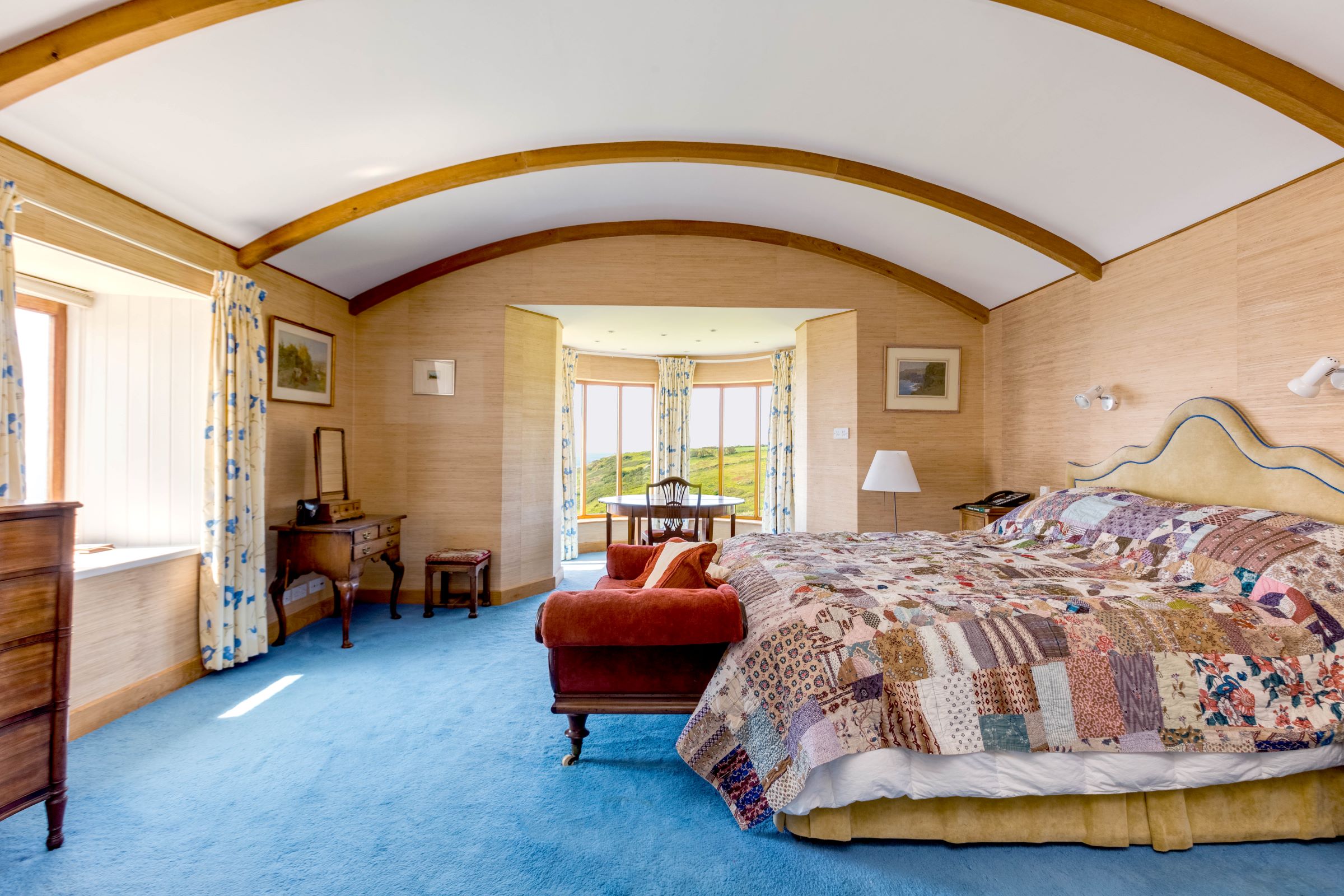 The master bedroom has a curved wooden beam ceiling and window sitting area and a table facing the window looking onto the sea