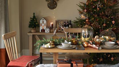 christmas dining table with foliage and house decorations in glass cloches with christmas tree behind