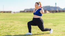 A woman wearing headphones performs a lunge in a park. She is holding a pair of dumbbells above her shoulders. 