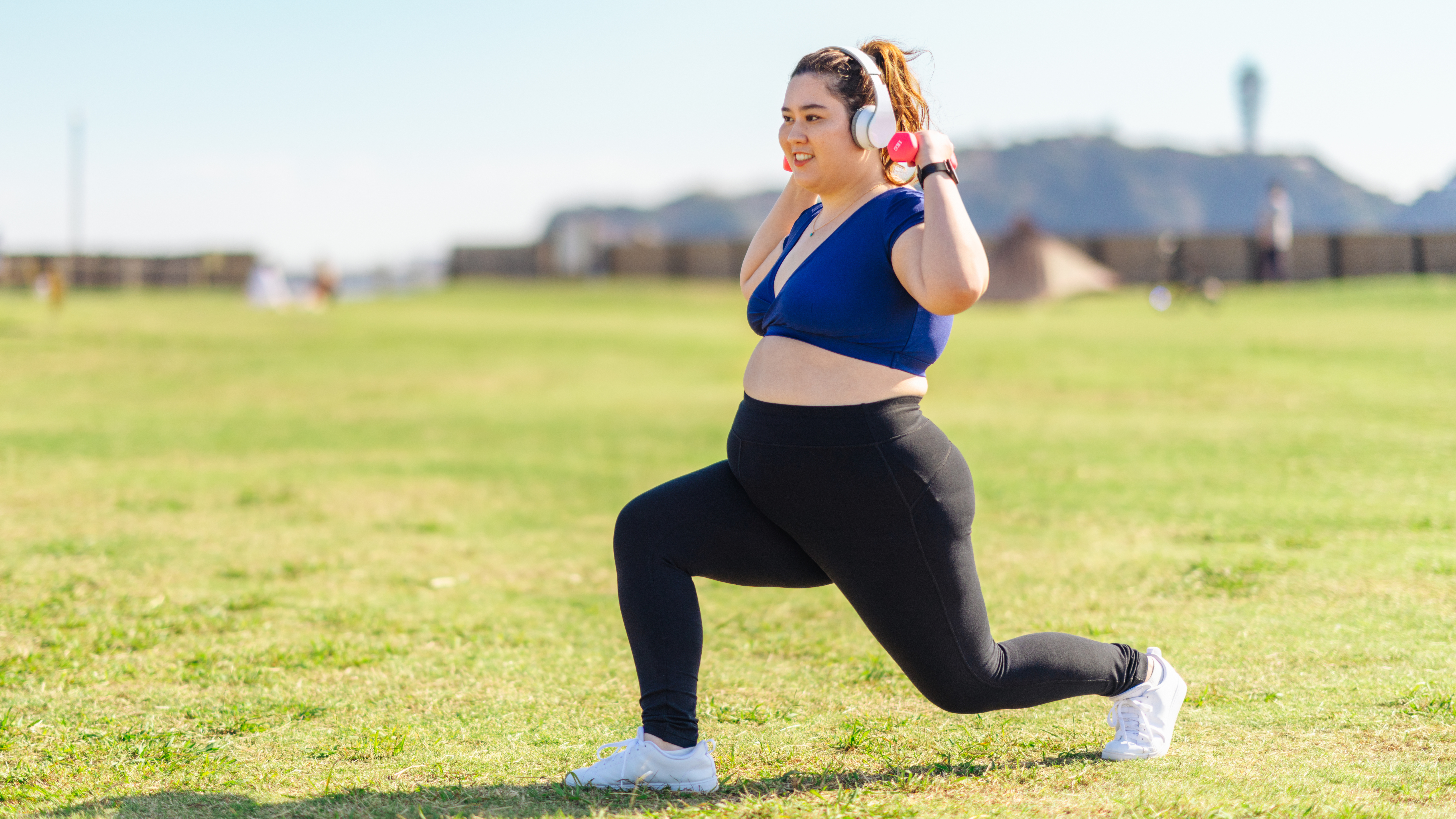 A woman wearing headphones performs a lunge in a park. She is holding a pair of dumbbells above her shoulders.
