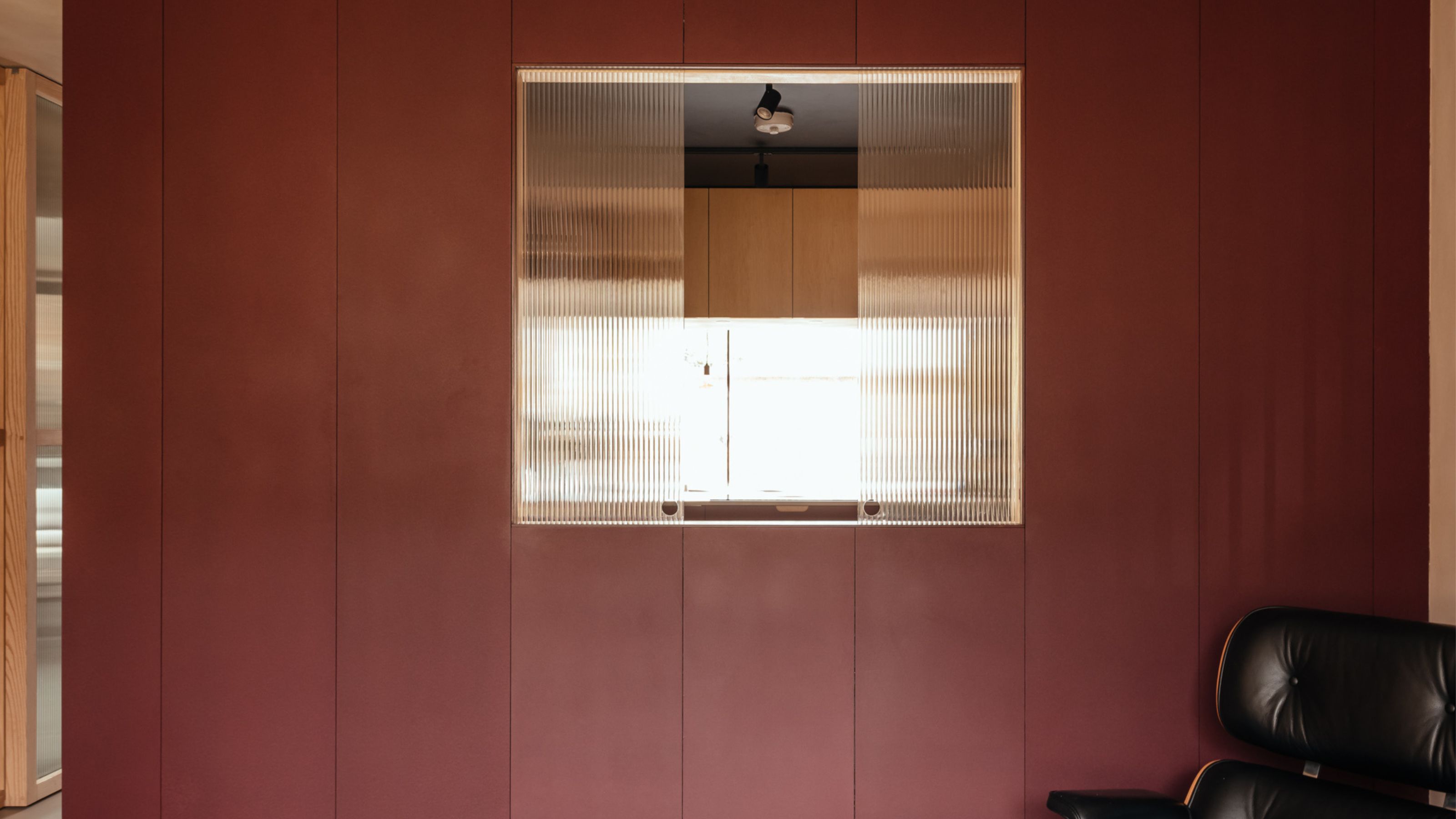 A kitchen with a red wall, a black chair and a reeded glass opening 