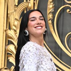 a woman with long, dark hair wearing a white, frilly top standing in front of gold doors