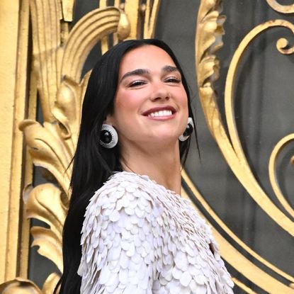 a woman with long, dark hair wearing a white, frilly top standing in front of gold doors