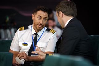 Colin (Ross Adams) sits in a bank of chairs with Piers (Geoffrey Breton), whose back is to the camera. Colin is dressed in his uniform and leaning in with a slightly cynical look on his face.