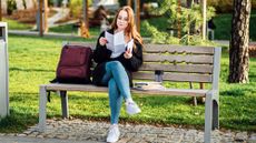 Young female student wearing glasses sitting outside on a bench next to her backback and books, reading a letter