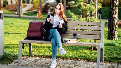 Young female student wearing glasses sitting outside on a bench next to her backback and books, reading a letter