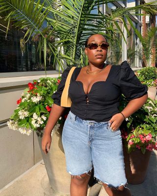 fashion and beauty editor Aniyah Morinia poses in front of pretty plants and flowers wearing tort oval sunglasses, a sweatheart top with puff sleeves, tan shoulder bag, and Bermuda denim cutoff shorts