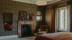 A bedroom with patterned wallpaper, a mirror, a fireplace, a soft sofa chair with a foot rest, and an overhead light