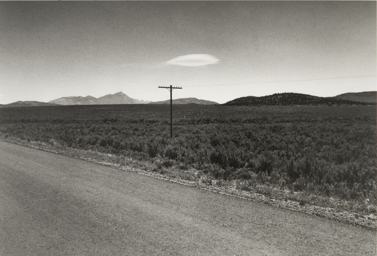 A black and white photograph of a deserted road running past scrubland toward distant mountains, with a telephone pole and a lens-shaped cloud overhead.